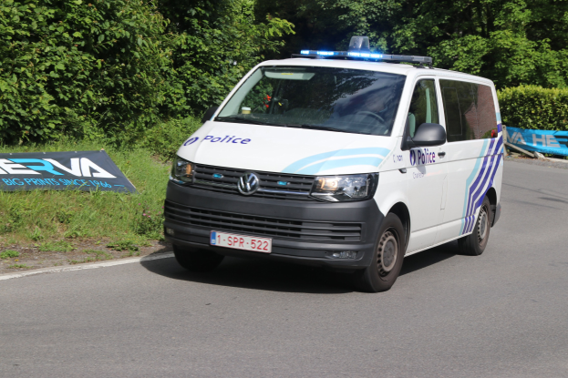 Ein weißer, blauer und schwarzer Polizeiwagen mit Beschriftung fährt an einem grasbewachsenen Bereich mit einem Schild vorbei, neben einer bewaldeten Straße.