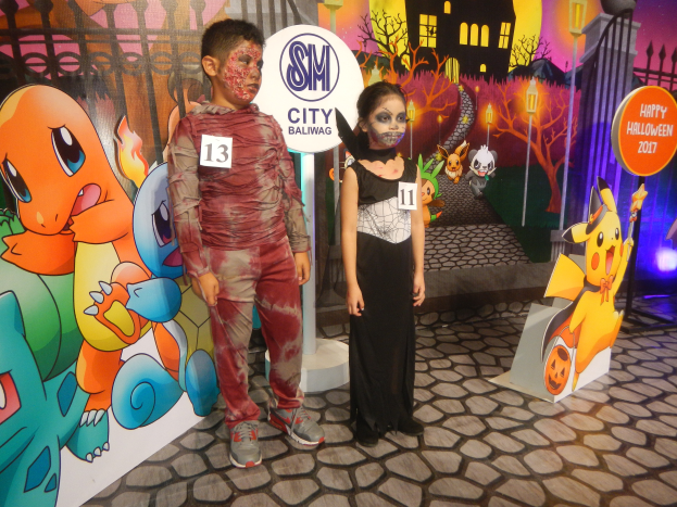 Zwei Kinder in Halloween-Kostümen stehen nebeneinander vor einer Wand mit einem Gemälde eines Gebäudes, Bäumen, Laternenmasten und einem Zaun, mit Brettern, die Text und Tierbilder zeigen, und festlicher Beleuchtung.