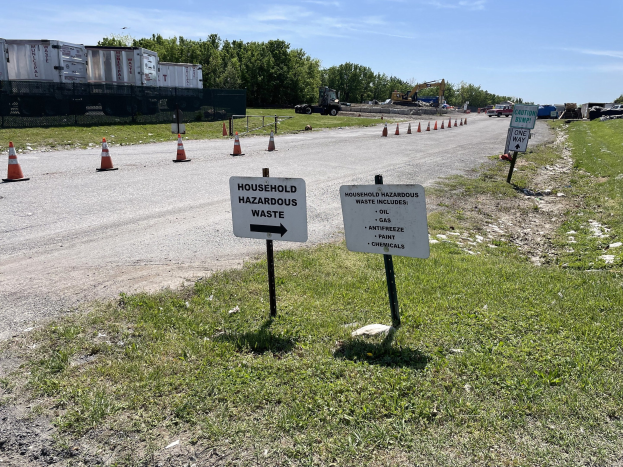 Straßensammelstelle für "Haushaltsgefährliche Abfälle" mit einem Schild markiert, umgeben von Gras, Verkehrskegeln, Fahrzeugen, Bäumen und einem bewölkten Himmel im Hintergrund.