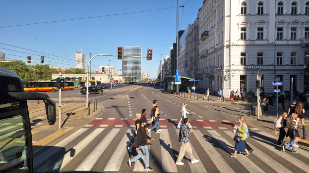 Gruppe von Menschen, die eine Straße an einer Ampel überqueren, mit einem Bus auf der linken Seite, Fahrzeugen auf der Straße, Verkehrssignalen, Laternenmasten, Gebäuden, Bäumen und einem klaren blauen Himmel.