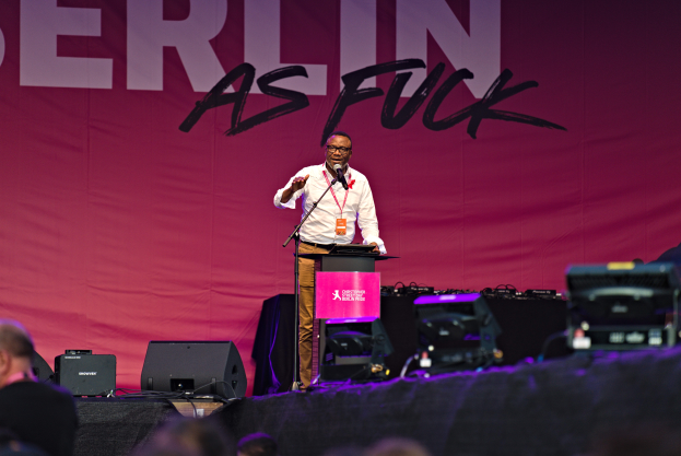 Ein Mann in weißem Hemd und braunen Hosen steht auf einer Bühne und hält ein Mikrofon in der Hand, hinter ihm eine rosa Banner mit der Aufschrift 'Berlin as Fuck' und Lautsprecher auf der Bühne.