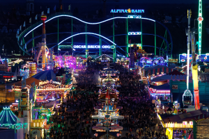 Eine große Menschenmenge, die um einen hell erleuchteten Karneval herumsteht, umgeben von Vergnügungsgeräten, Pfosten, Flaggen und Namensschildern, mit einem dunklen Hintergrund, der eine festliche Atmosphäre schafft.