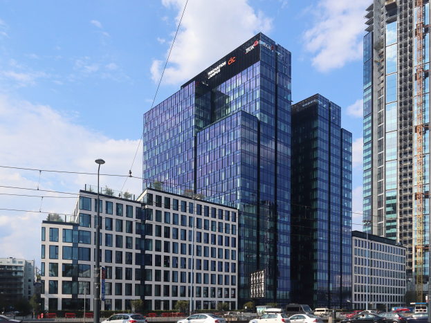 Hauptsitz der Deutschen Bank in Frankfurt, Deutschland, ein großes mehrstöckiges Gebäude mit zahlreichen Fenstern, umgeben von städtischer Infrastruktur einschließlich Straßenlaternen, Straßenlaternen, Strommasten, Stromkabeln, Kraftfahrzeugen auf der Straße, Büschen, Bäumen und einem Himmel mit Wolken im Hintergrund.