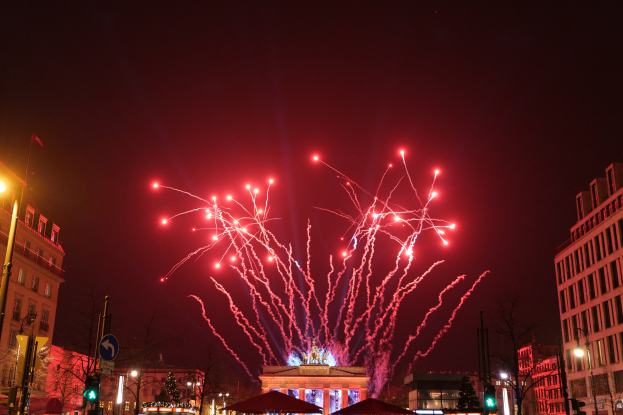 Eine belebte Straße in einer Stadt bei Nacht an Silvester in Berlin, mit Gebäuden, Bäumen, Laternenmästen, Ampeln, Schildern, Zelten, Menschen und einem prächtigen Feuerwerk am Himmel.