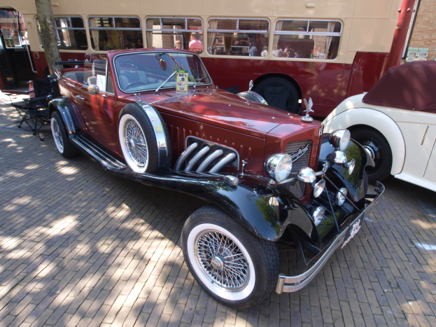 Ein Oldtimer neben einem Doppeldeckerbus auf einem Kopfsteinpflasterstraßen, mit Stühlen und einem Baum im Vordergrund, einem Gebäude mit Fenstern im Hintergrund und Menschen im Bus zu sehen.