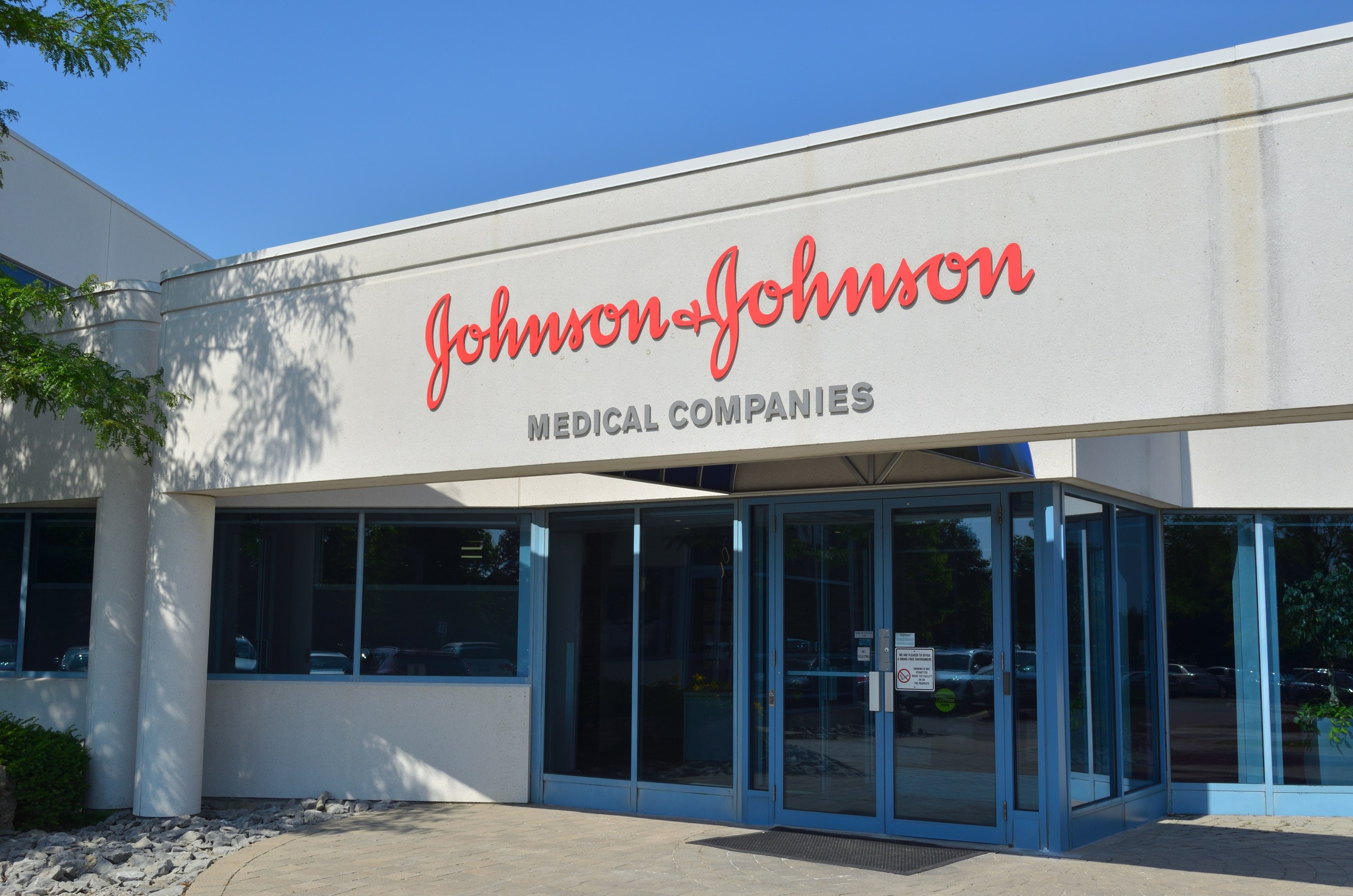 Außensicht des Johnson & Johnson Medical Companies Gebäudes mit Glaswänden, Text an der Fassade, Steinen, Pflanzen und einem Baum auf der linken Seite, unter einem klaren blauen Himmel.