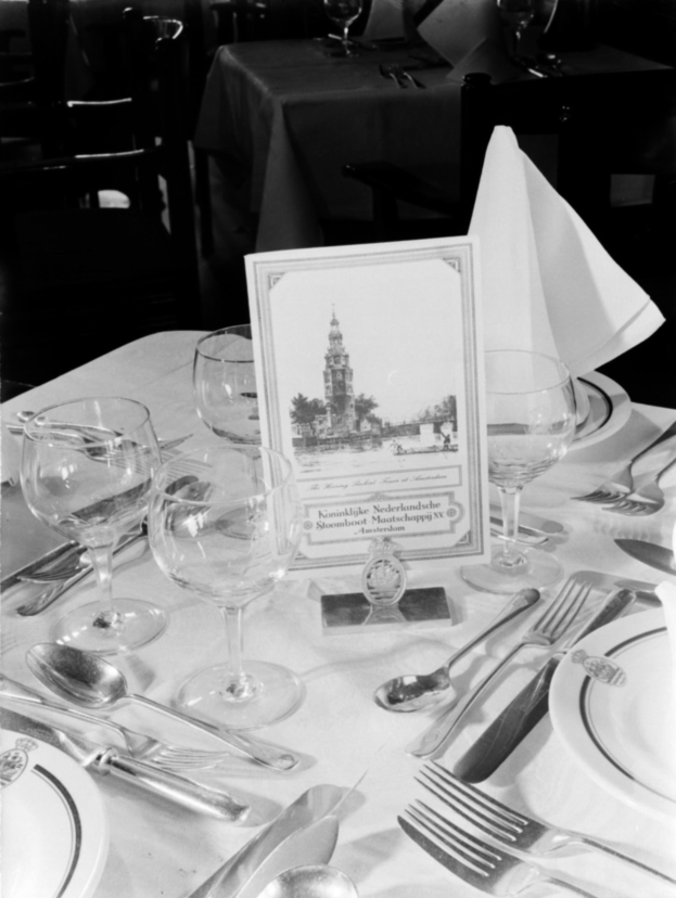 Ein Schwarz-Weiß-Foto eines festlich gedeckten Banketttisches mit Tellern, Besteck, Gläsern und Servietten, mit anderen Tischen und Stühlen im Hintergrund.