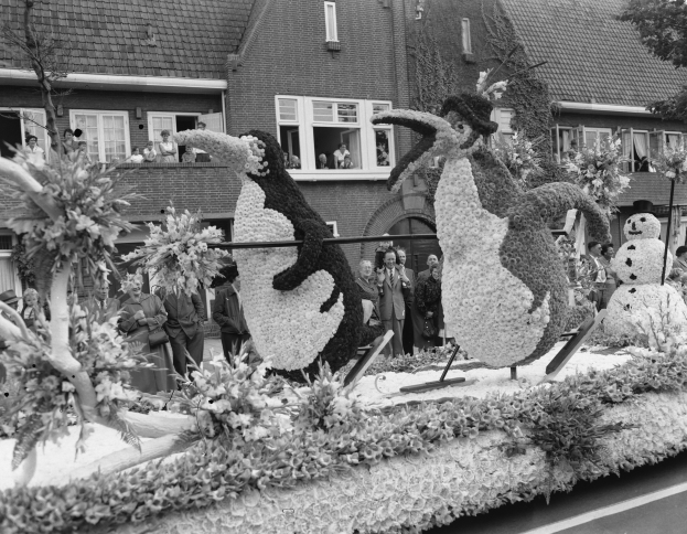 Schwarzes und weißes Foto eines Paradewagen mit Blumen und Pinguinen, mit Menschen, Bäumen und Gebäuden im Hintergrund.