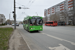 Ein grüner Bus fährt auf einer von hohen Gebäuden gesäumten Straße, mit Gras und Strommasten auf der linken Seite und Bäumen vor einem klaren blauen Himmel im Hintergrund.