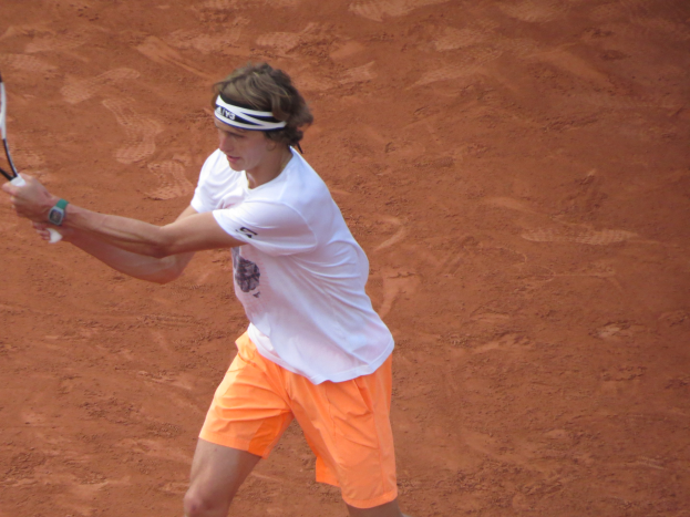 Ein Mann in einem weißen T-Shirt und orangefarbenen Shorts hält einen Tennis-Schläger auf einem Sandplatz, bereit, einen Ball zu schlagen.
