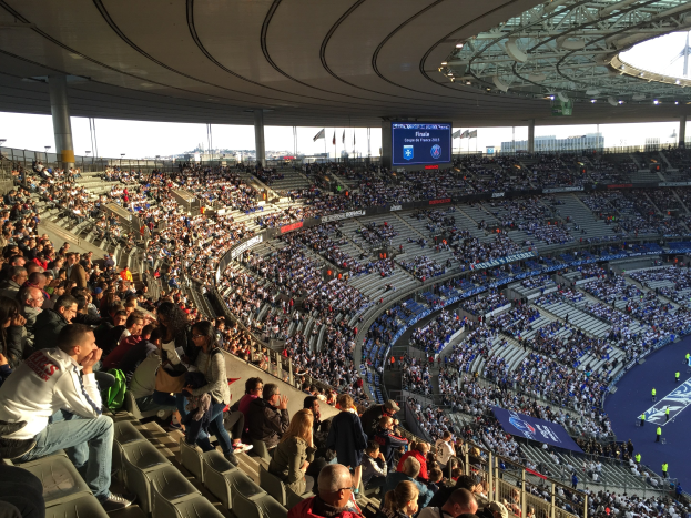 Large crowd in a stadium watching a soccer game, with a stage, flags, poles, and a screen visible in the background.