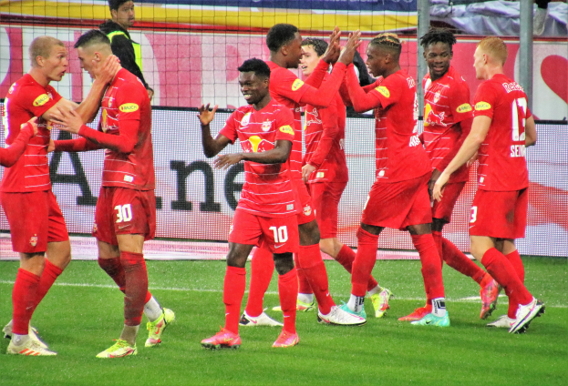 Eine Gruppe von Fußballspielern feiert ein Tor auf einem grünen Feld, mit einem Netz, Bannern mit Text, Pfosten und anderen Gegenständen im Hintergrund.