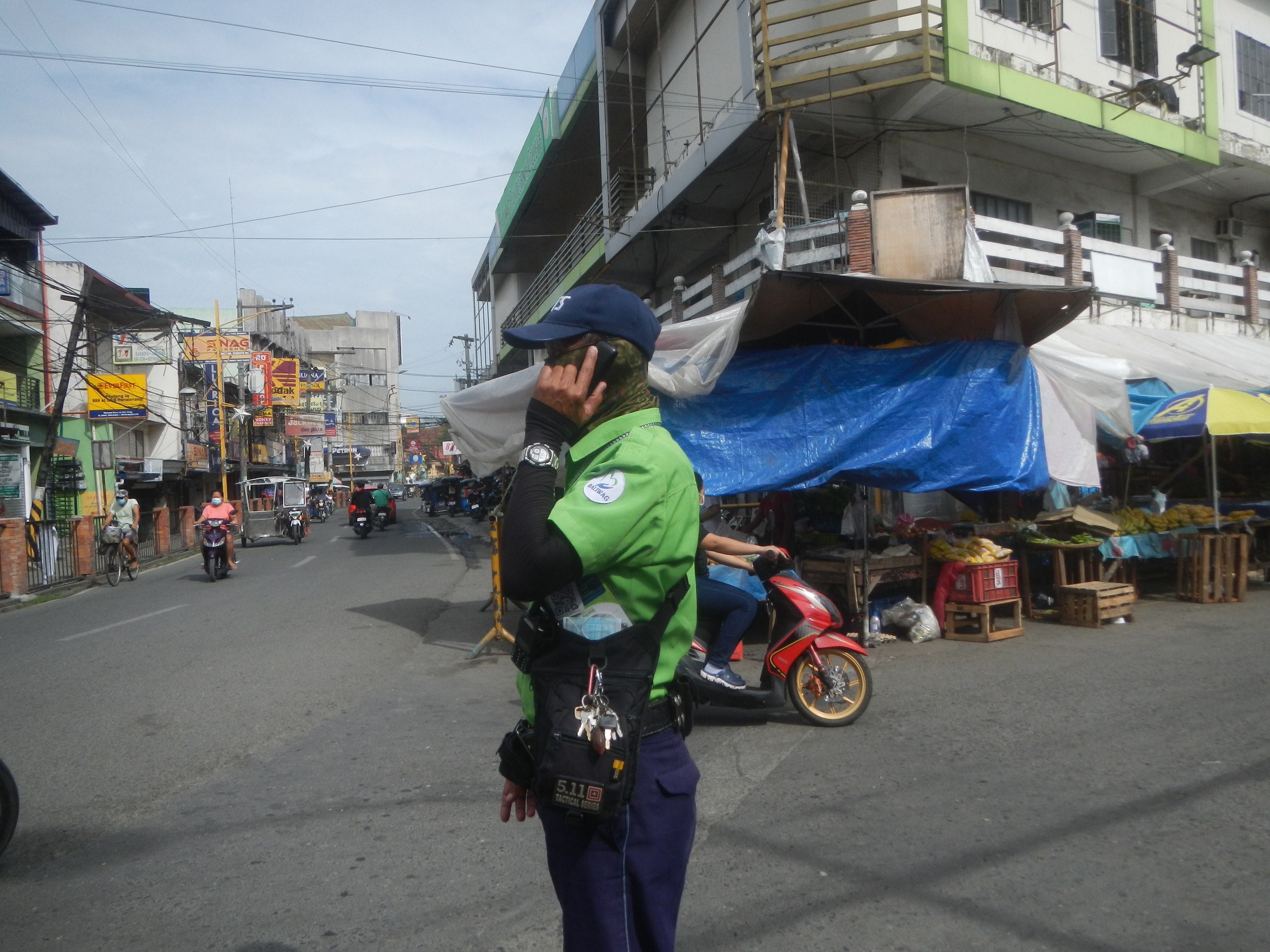 Ein Polizeibeamter in grüner und blauer Uniform mit blauer Mütze steht auf einer Straße, spricht auf einem Handy, mit Fahrzeugen und Marktbuden hinter ihm und einem Gebäude im bewölkten Himmel Hintergrund.
