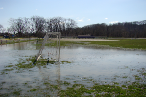 Ein Fußballtor steht inmitten eines überfluteten Feldes, umgeben von Gras, einem Zaun, Bäumen, Häusern und unter einem bewölkten Himmel.