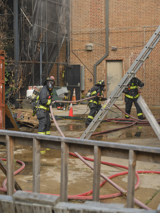 Feuerwehrleute mit Helmen arbeiten daran, ein Gebäude Feuer zu löschen, umgeben von Equipment und einem Metallzaun.