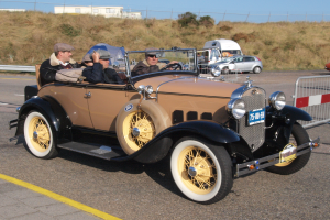 Eine Gruppe von Menschen in einem Oldtimer auf einer Straße mit einer Begrenzung auf der rechten Seite, andere Fahrzeuge, Bäume, Gebäude und einen klaren blauen Himmel im Hintergrund.