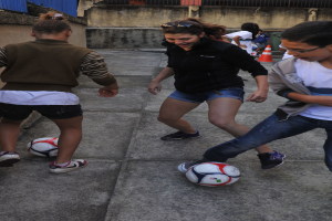 Eine Gruppe junger Menschen, die auf einem Bürgersteig Fußball spielen, mit einer Wand im Hintergrund, einem Verkehrskegel im Vordergrund und einem Gebäude in der Ferne, alle mit Schuhen bekleidet.