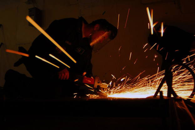 Zwei Männer schweißen ein Metallstück in einem dunklen Raum, Funken sprühen von der Schweißmaschine, mit einer Wand im Hintergrund.