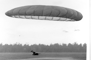 Schwarzes und weißes Foto einer Person, die mit einem Gleitschirm fliegt und von Bäumen und einem klaren Himmel umgeben ist, mit Text unten.