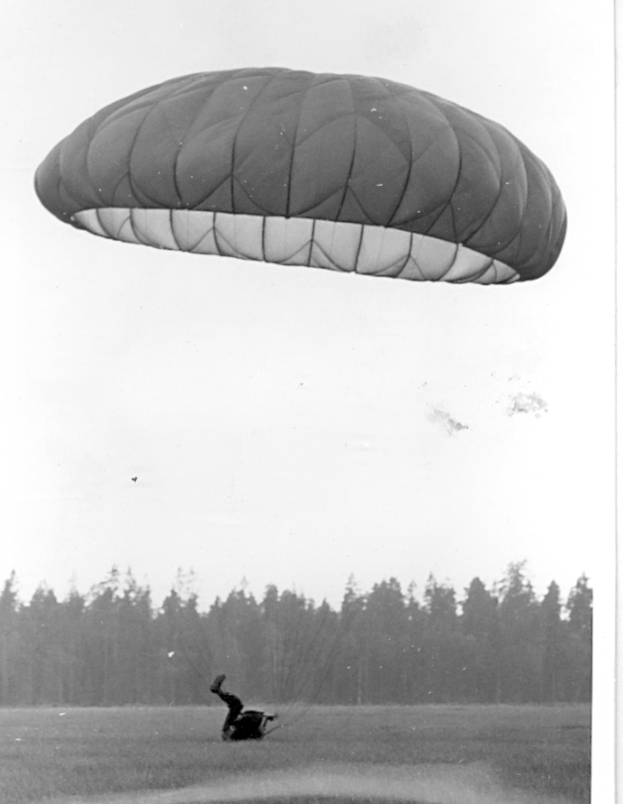 Schwarzes und weißes Foto einer Person, die mit einem Gleitschirm fliegt und von Bäumen und einem klaren Himmel umgeben ist, mit Text unten.