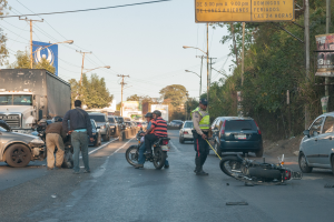 Eine Gruppe von Menschen steht um ein verunglücktes Motorrad auf der Straße mit mehreren Fahrzeugen, darunter ein Lastwagen, und einem Hintergrund aus Bäumen, Pfosten, Lampen, Schildern und Himmel.