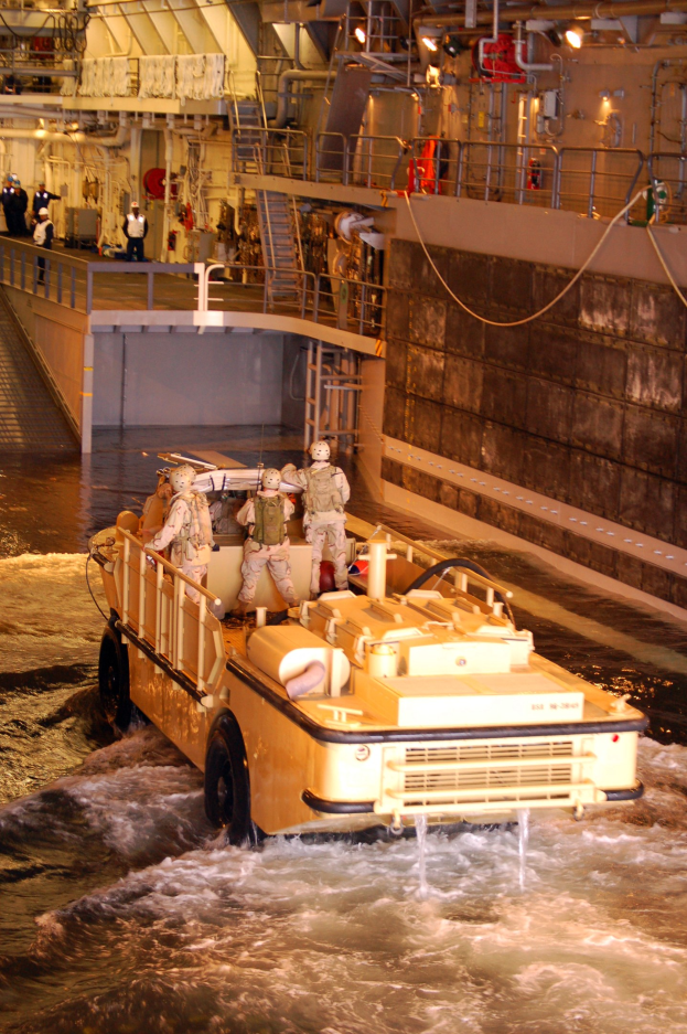 Amphibisches Fahrzeug fährt durch Wasser mit Helmträgern an Bord, neben einer Wand und Hintergrundstrukturen einschließlich Geländern, Lampen und Rohren.