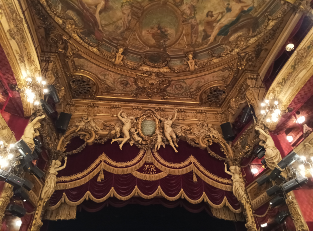 Theaterinnenraum mit roter Gardine, Kronleuchtern, Deckenplastiken, Lichtern und einem Gemälde, das eine Bühne und eine kleine Gruppe von Menschen vorne zeigt.