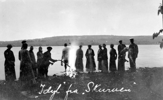 Gruppe von Menschen um ein Feuer am Ufer eines Sees mit Bäumen im Hintergrund und einem klaren Himmel, schwarz-weißes Bild mit Text unten.