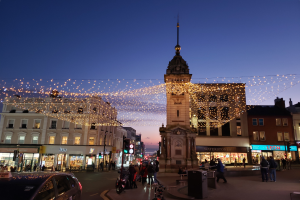 Eine belebte Stadtstraße bei Nacht mit Menschen, Fahrzeugen, Fahrrädern, Mülltonnen, Gebäuden, Laternen, Ampeln und einem Uhrenturm im Hintergrund, beleuchtet von Weihnachtslichtern.
