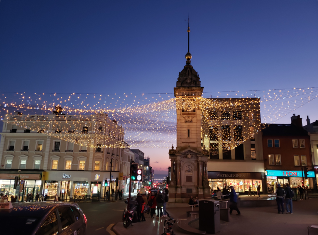 Eine belebte Stadtstraße bei Nacht mit Menschen, Fahrzeugen, Fahrrädern, Mülltonnen, Gebäuden, Laternen, Ampeln und einem Uhrenturm im Hintergrund, beleuchtet von Weihnachtslichtern.