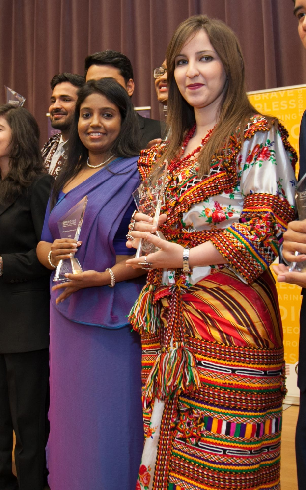 Gruppe von Menschen auf einer Bühne mit einer Frau in der Mitte, die eine Trophäe hält, dahinter ein Vorhang und eine Tafel mit der Aufschrift "Bravo Segen Award Gewinner."