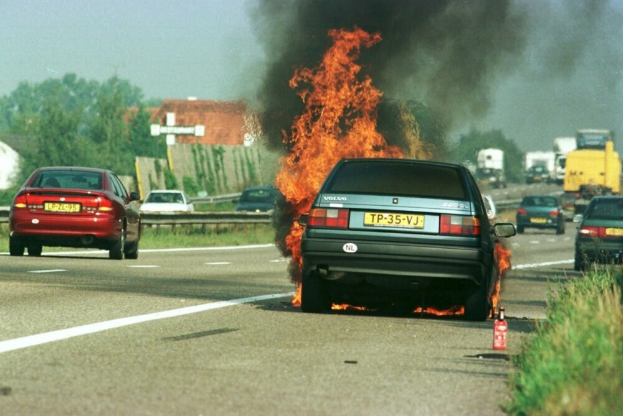 Ein Auto in Flammen am Straßenrand mit anderen Fahrzeugen in der Nähe, Bäumen, Gebäuden und einem klaren blauen Himmel im Hintergrund und Gras mit einem Feuerlöscher auf der rechten Seite.
