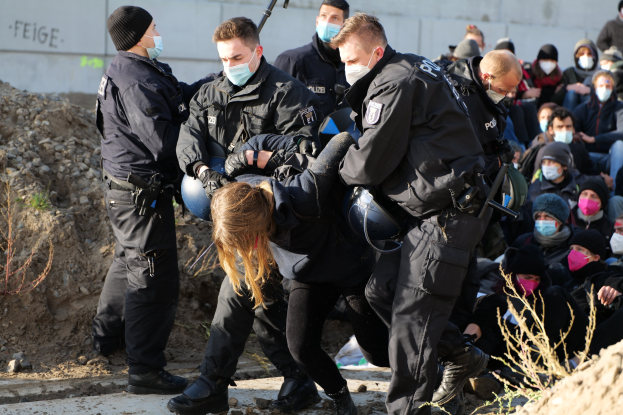 Eine Gruppe maskierter Menschen in Mützen, einige mit Waffen, umringen eine Frau, die von der Polizei eskortiert wird, mit Pflanzen, Steinen und einer Wand mit Protesttext im Hintergrund.