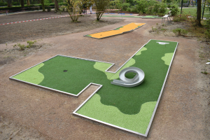 Minigolfanlage in einem Park mit Grünflächen, Menschen, Gebäuden und einem klaren Himmel im Hintergrund.