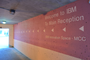 Eine Wand mit 'Willkommen bei IBM' beleuchtet von Deckenlampen mit einer Tür zum IBM Innovation Space MCC im Hintergrund.