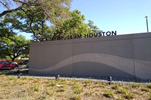 Außenansicht des Holocaust-Museums Houston mit eingravierten Textwänden, verstreuten Steinen, Grünflächen, Bäumen, vorbeifahrenden Fahrzeugen, einer Straßenlaterne und einem bewölkten Himmel.