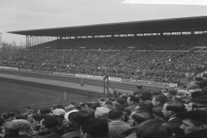 Schwarzes und weißes Foto einer vollen Stadion mit Zuschauern, die ein Fußballspiel verfolgen, mit Bannern, Pfosten, einer Hütte, Bäumen, einem Turm und einem bewölkten Himmel.