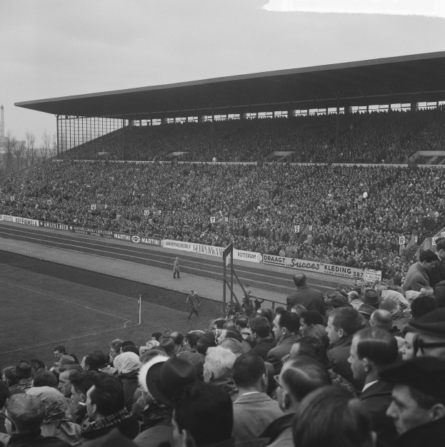 Schwarzes und weißes Foto einer vollen Stadion mit Zuschauern, die ein Fußballspiel verfolgen, mit Bannern, Pfosten, einer Hütte, Bäumen, einem Turm und einem bewölkten Himmel.