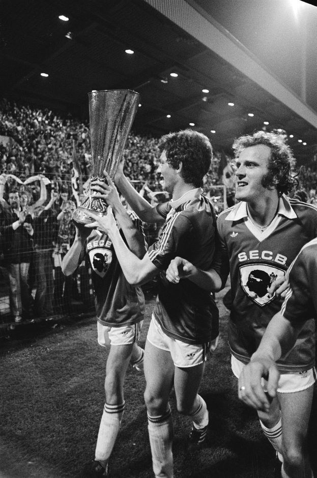 Eine Gruppe von Männern, die einen Pokal auf einem Fußballfeld halten und ihren Sieg im FA Cup Final feiern, umgeben von einer Menge hinter einem Zaun, unter Stadionbeleuchtung, in Schwarz-Weiß.