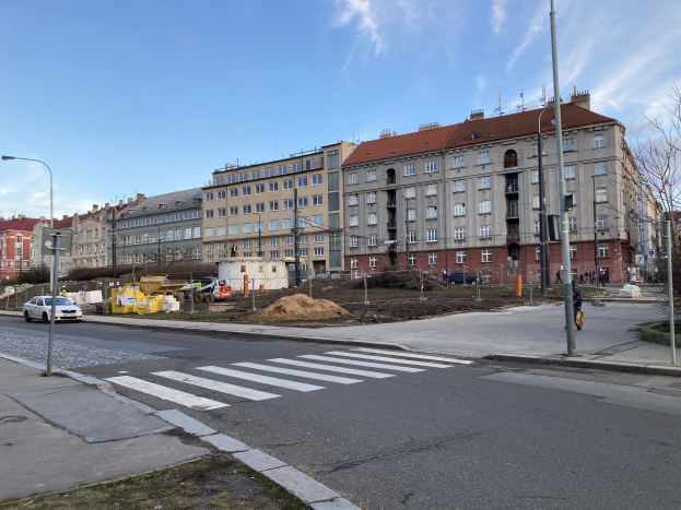 Baustelle für ein neues Wohnhaus im Vordergrund einer städtischen Straße mit parkenden Autos, Laternen, Bäumen, Gebäuden mit Fenstern und einem bewölkten Himmel.