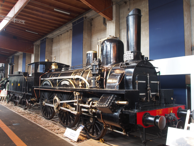 Eine große, alte Dampflokomotive, die auf Eisenbahnschienen in einem Museum ausgestellt ist, umgeben von Informationsschildern und Ständen, mit einer beleuchteten Decke und Wand im Hintergrund.