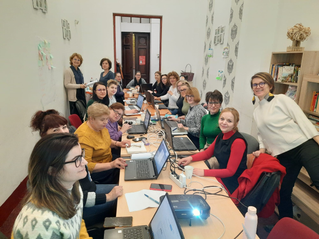 Eine Gruppe von Frauen mit Behinderungen nimmt an einem Workshop teil, sitzt um einen Tisch mit Laptops, Papieren und anderen Gegenständen, mit Jacken auf Stühlen, einem Bücherregal und Papieren an der Wand.