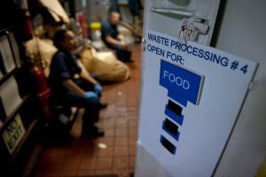 Ein Mann sitzt auf dem Boden neben einem Schild mit der Aufschrift "Abfallverarbeitung 4: Offen für Lebensmittel", mit einer Tür auf der rechten Seite, Regalen mit Gegenständen auf der linken Seite und unscharfen Figuren und Taschen im Hintergrund.