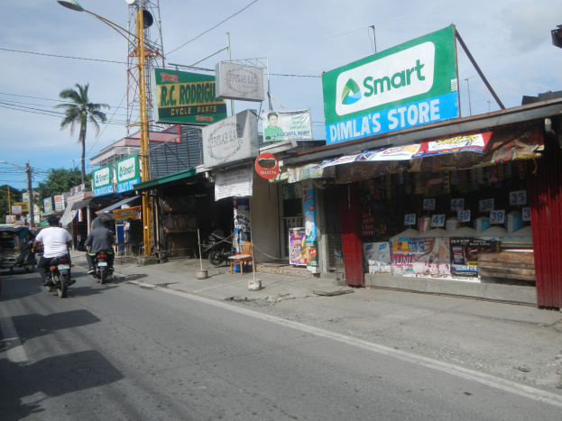 Eine belebte Straße mit Motorradfahrern, Autos, Gebäuden, Schildern, Strommästen, Drähten, Straßenlaternen, Bäumen und einem bewölktem Himmel.