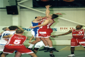 Eine Gruppe von Männern, die Basketball auf einem Court mit einer Wandmontage und Text im Hintergrund spielen, mit einem Basketball in der Luft darüber.