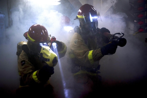 Zwei Feuerwehrleute in Schutzausrüstung sprühen Wasser auf eine rauchgefüllte Wand, mit einer Tafel und Gegenständen auf beiden Seiten.