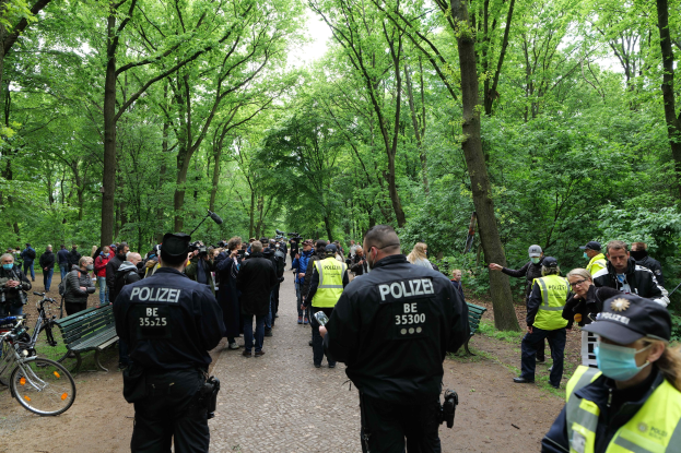 Polizisten vor einer Menschenmenge während einer Outdoor-Veranstaltung, mit Fahrrädern und einer Bank im Vordergrund und Bäumen im Hintergrund.