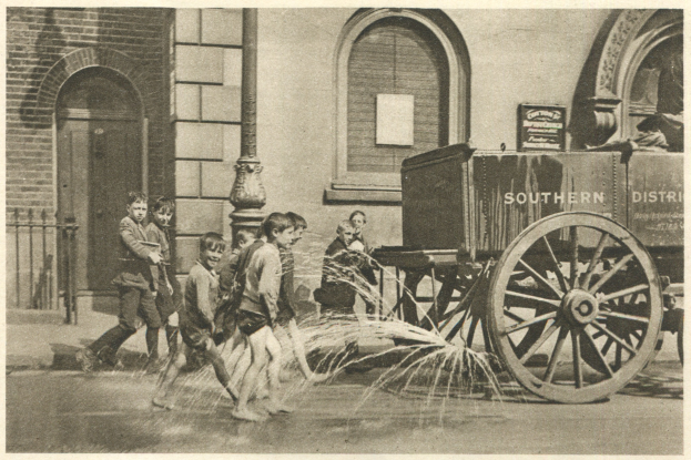 Schwarzes und wei├čes Foto von Kindern, die mit einem Feuerhydranten vor einem Geb├Ąude mit Fenstern, einer Tür, einem Mast und einem Zaun spielen, mit einem Wagen im Hintergrund.