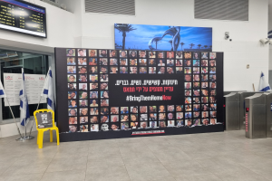 Eine große Wand zeigt mehrere Bilder, einen gelben Stuhl, israelische Regierungsflaggen auf Stangen, eine Tafel mit dem Text 'Bring Them Home Now', einen montierten Bildschirm, Deckenleuchten und andere Gegenstände.