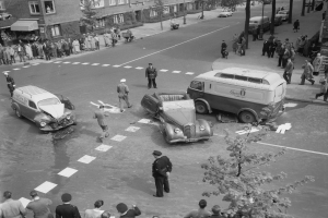 Schwarz-weiß-Szene eines Autounfalls an einer Straßenkreuzung mit einem Lieferwagen, anderen Fahrzeugen und einer Gruppe von Menschen in der Nähe, mit Gebäuden, Bäumen, Pfählen und Texttafeln im Hintergrund.